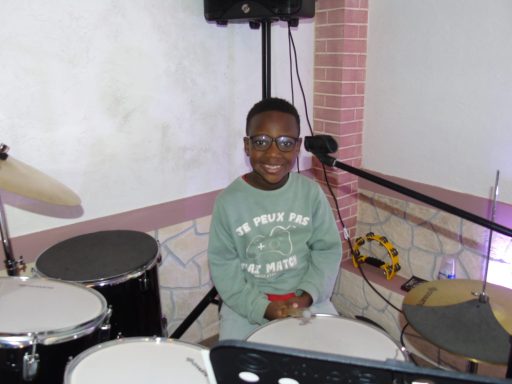 Un enfant souriant assis derrière une batterie dans une salle de répétition.