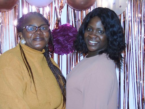 Deux femmes souriantes devant un fond décoratif avec des ballons et des éléments festifs.
