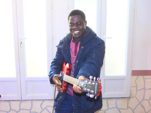 Homme souriant tenant une guitare, portant un manteau bleu.